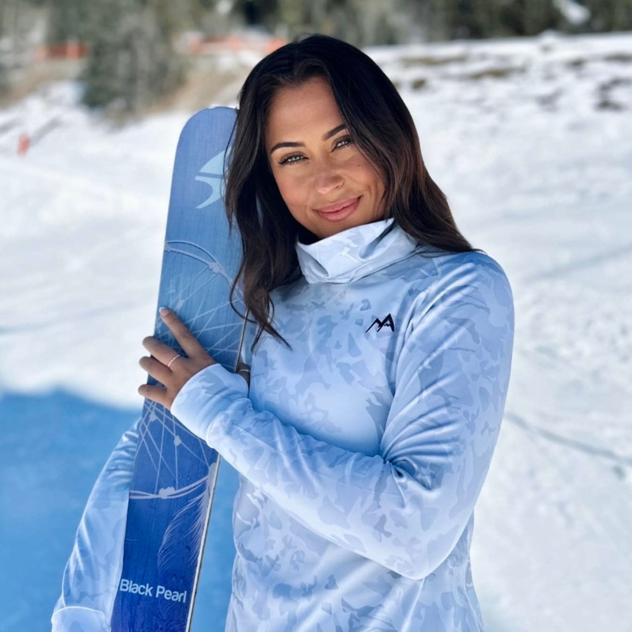 Model standing in the snow holding skis wearing Arctic Eco-Sno Women's Base Layer Top in Sno-Camo.