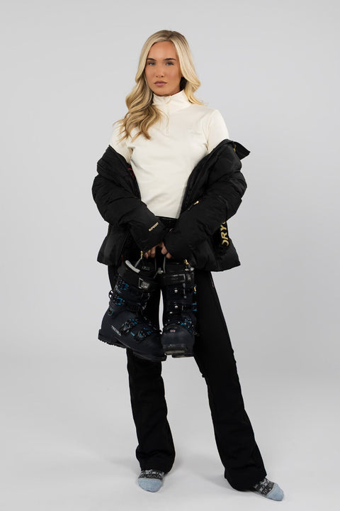 Woman wearing Arctic Eco-Sno women's Buttercream quarter zip thermal ski base layer top under an open black ski jacket, holding ski boots with black ski trousers.