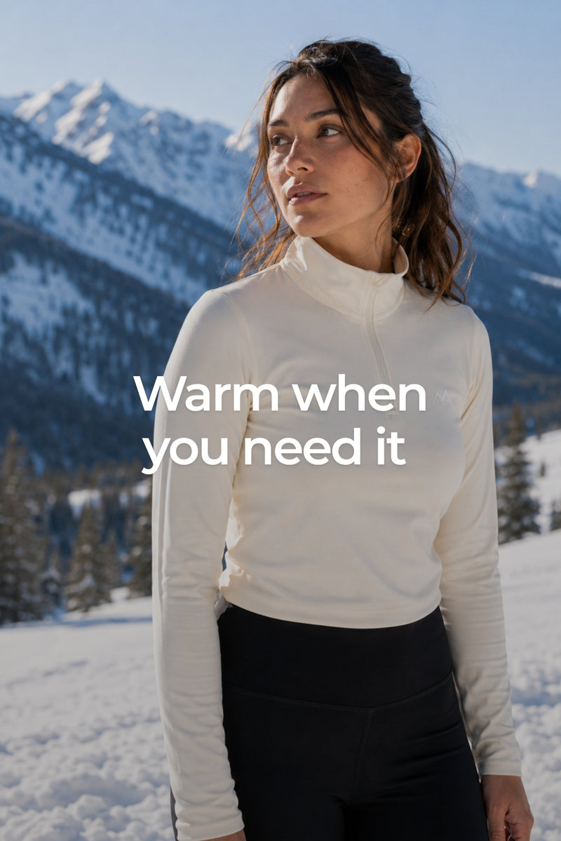 A woman standing in a snowy mountain setting wearing a fitted butter cream long-sleeve quarter-zip base layer top and black leggings, with the text 'Warm when you need it' overlaid across the center.