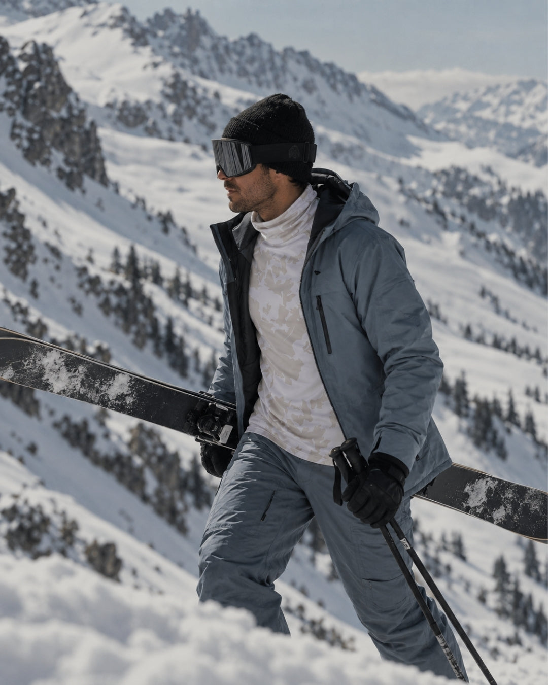 A man in ski gear, including a slate blue jacket, matching pants, a white turtleneck, black beanie, and goggles, stands in the snow holding skis on a mountain slope.