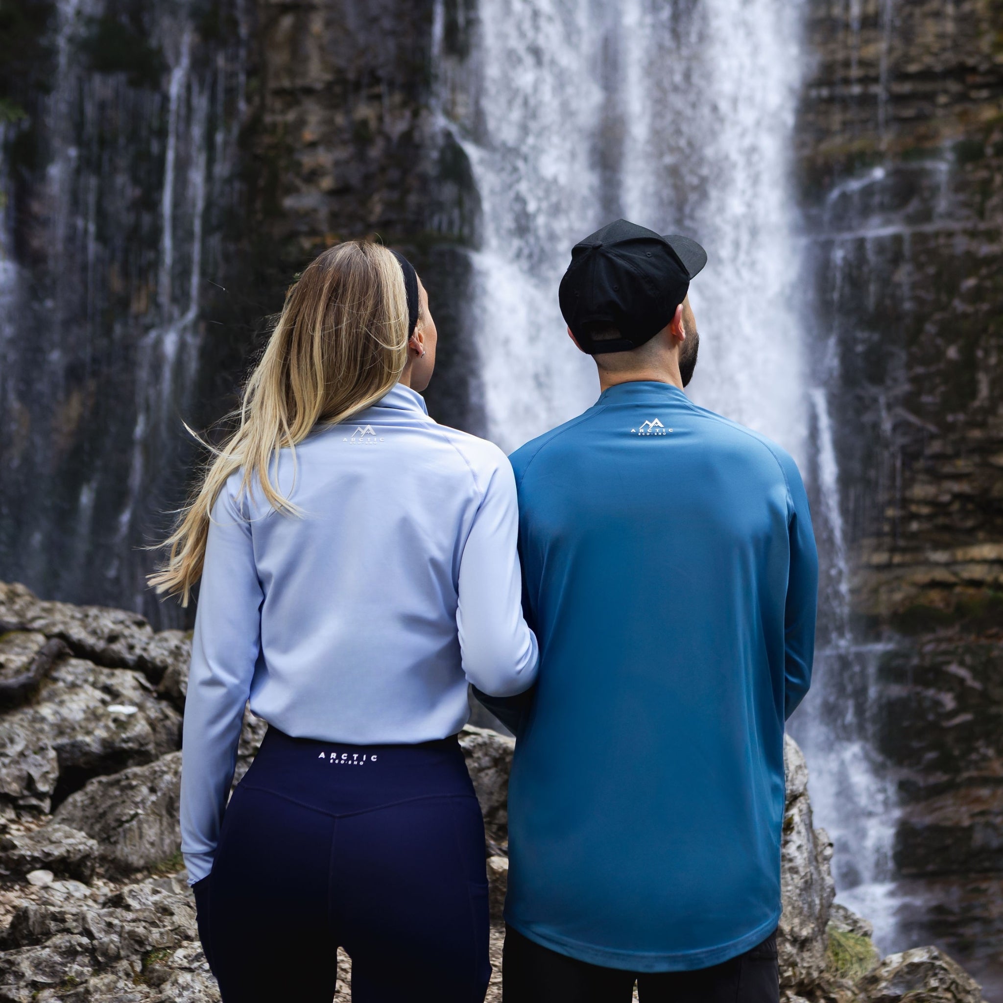 Two people wearing arctic eco-sno base layers standing in front of a waterfall.